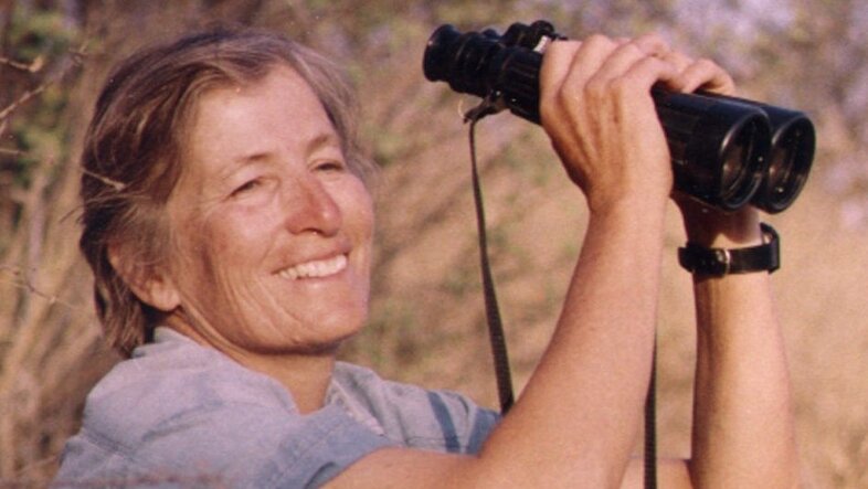 Photo of Katy Payne in the savannah, sitting with a binocular in her hands and smiling