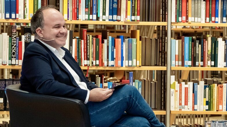 Porträtfoto von Gerold Riedmann, sitzend vor Bücherregal 