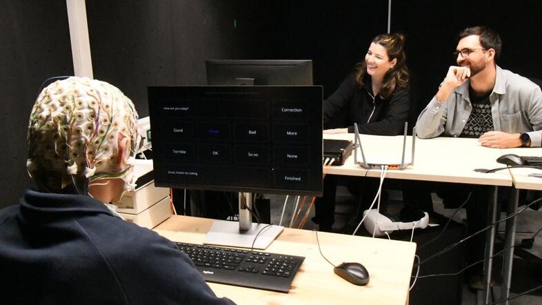 Eine Person mit EEG-Messhaube sitzt zwei Personen, die das Experiment leiten, gegenüberter Experiment 