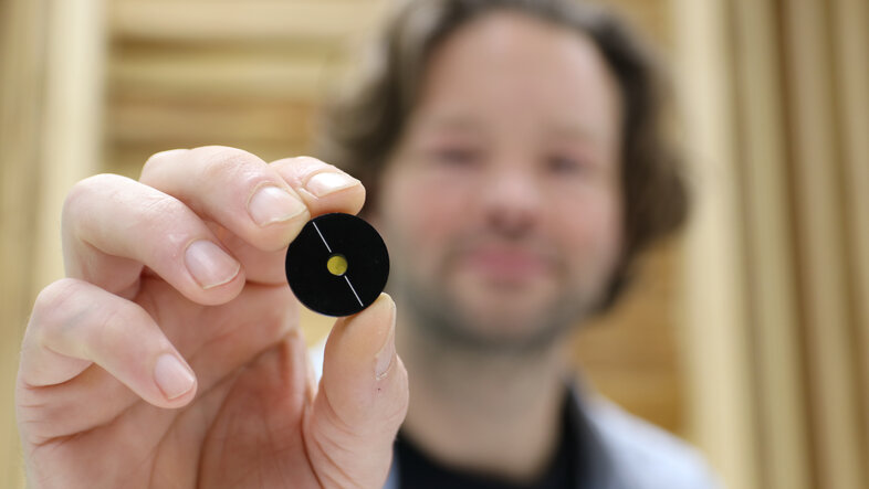 Lee Rozema (blurred) hold up a small black disk with a transparent yellow centre.