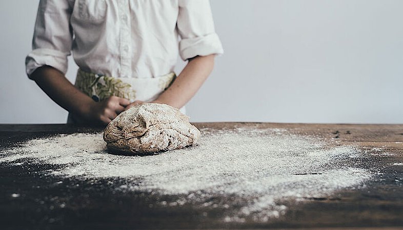 Der Teig für einen Laib Brot liegt auf einer mit Mehl bestäubten Arbeitsfläche und im Hintergrund steht eine Person mit Bäckerjacke und Schürze