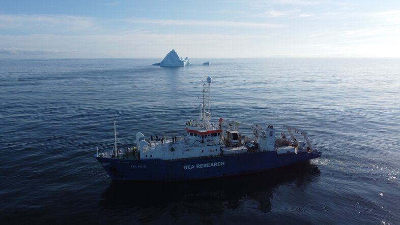 Research vessel "SEA RESEARCH" sails in open ocean with a distant iceberg under cloudy skies.