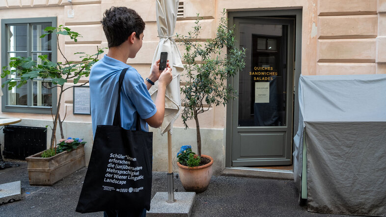 Schüler mit einem Totebag fotografiert mit seinem Handy die Glastür eines Lokals