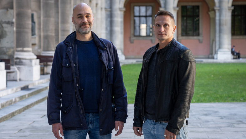 Zwei Herren um die 40 stehen nebeneinander im Innenhof des Hauptgebäudes der Universität Wien 