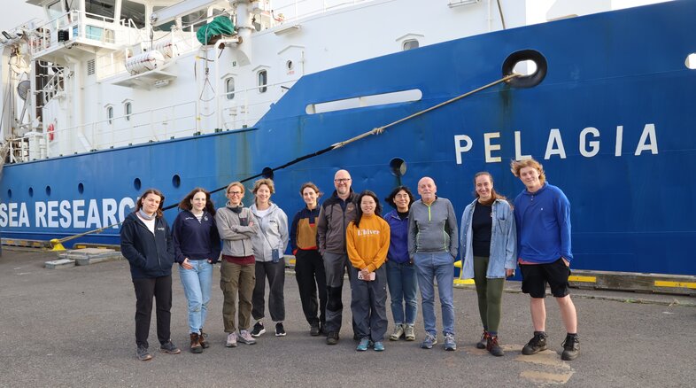 Ten people pose in front of blue research vessel "PELAGIA" with "SEA RESEARCH" on its side.