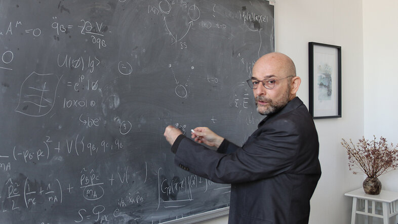 Brukner steht vor einer mit mathematischen Formeln beschriebenen Tafel und dreht sich zum Fotografen um. Im Hintergrund ein Wandbild und ein Blumenstrauß auf einem Podest.