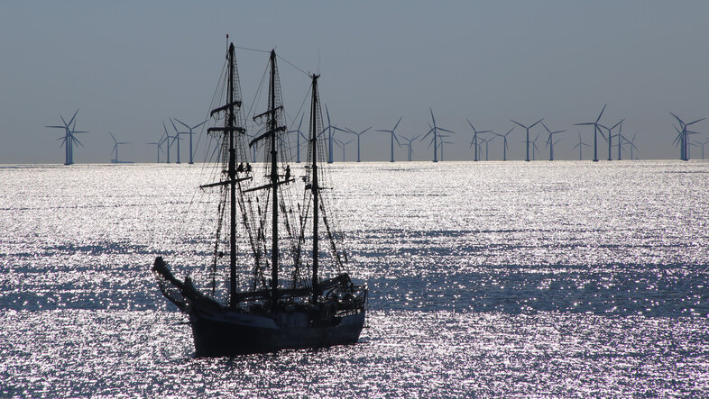 © Ein abgetakeltes Segelschiff auf ruhiger, sonnenglitzernder See. Im Hintergrund ein Windpark.