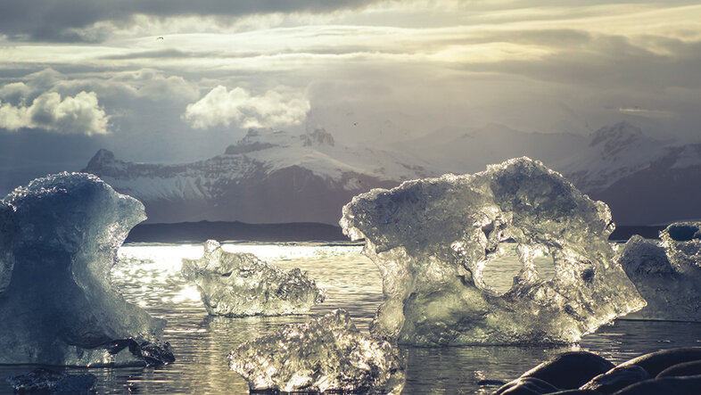 Schmelzende, bereits durchsichtige Eisblöcke im sonnenglitzernden Meer. Im Hintergrund schneebedeckte Berge.
