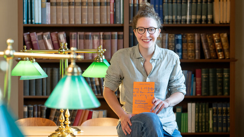 Die Forscherin Natascha Bobrowsky sitzt für das Fotoshooting locker auf dem Tisch in der Fachbereichsbibliothek Geschichte, ihr Buch hat sie in der Hand
