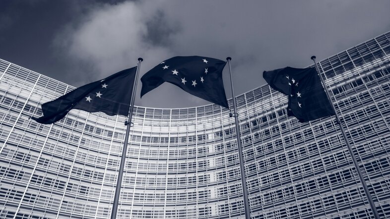Drei EU-Flaggen in der Froschperspektive vor dem EU-Parlament, dunkle Farbtöne