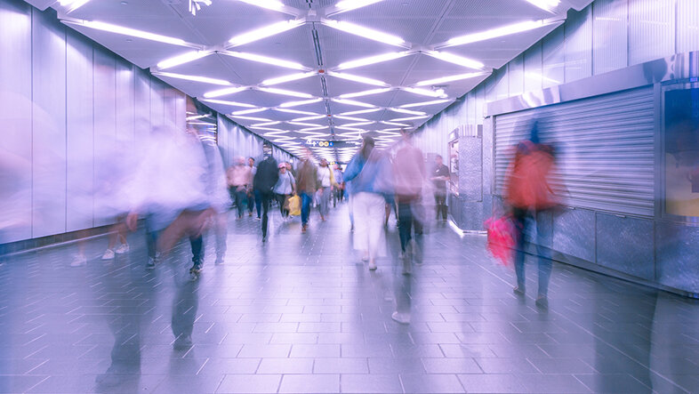 Bewegt verschwommene Menschen in einer U-Bahn-Passage. Auf der rechten Seite bewegen sie sich ins Bild hinein, auf der linken Seite aus dem Bild heraus.