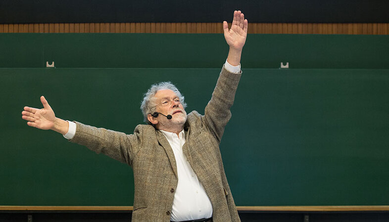 Anton Zeilinger in front of a green board in the lecture hall describes with his hands what he is explaining (raised arms)