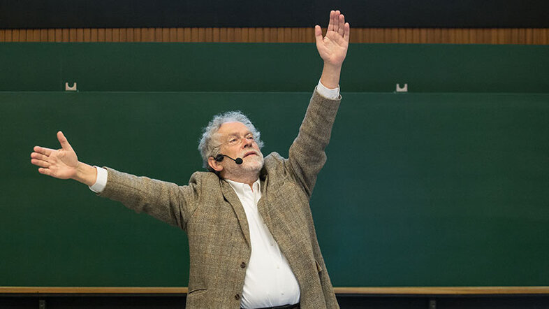Anton Zeilinger vor einer grünen Tafel im Hörsaal beschreibt mit den Händen, was er gerade erklärt (erhobene Arme)