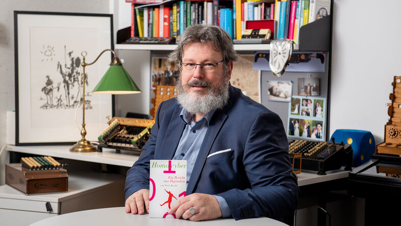 Porträtfoto von Peter Reichl am Schreibtisch, in der Hand sein neues Buch, im Hintergrund alte Rechenmaschinen