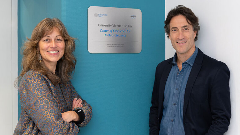 Manuela Schmidt und David Gómez Varela posieren vor einem Schild mit der Aufschrift "University of Vienna - Bruker Center of Excellence for Metaproteomics"