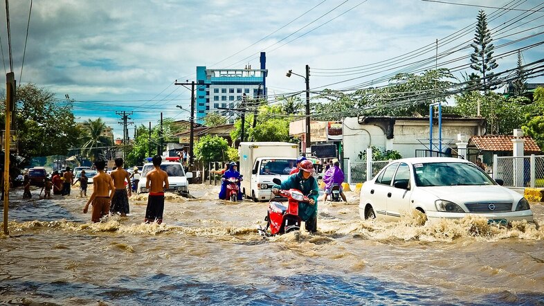 Überflutete Landschaft. Autos, Bäume und Gebäude stehen im Wasser, Menschen kämpfen sich durch die Wassermassen