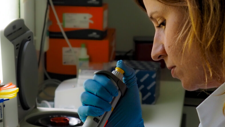 eine blonde Forscherin im Labormantel pipettiert eine klare Flüssigkeit in ein 50ml Reagenzglas