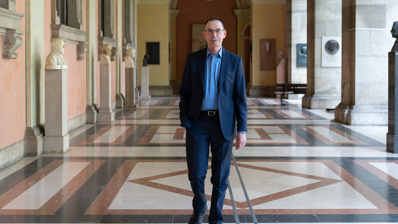 A man in a suit walks straight on towards the viewer in a hallway