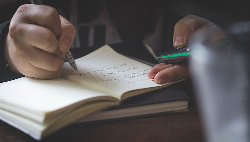 Eine Hand schreibt mit einem Stift in einem Notizbuch, während die andere ein Mobiltelefon hält.